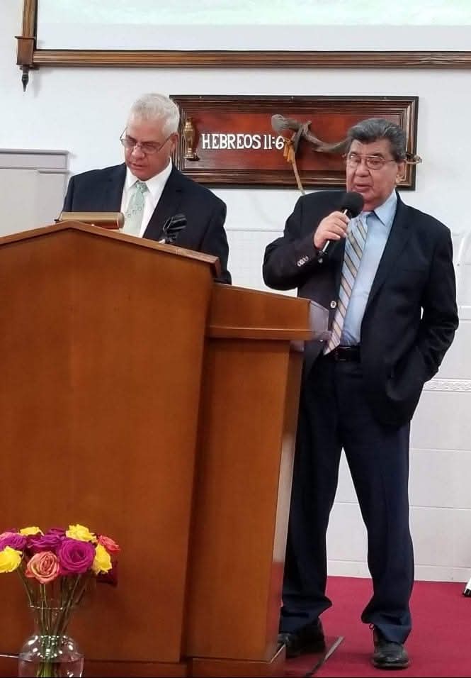 Two ministers at the wooden pulpit beneath the Hebrews 11:6 plaque