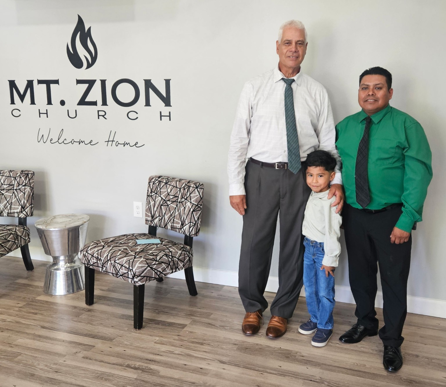 Pastor Jose Alvarado with a father and son in front of the Mt. Zion Welcome Home wall